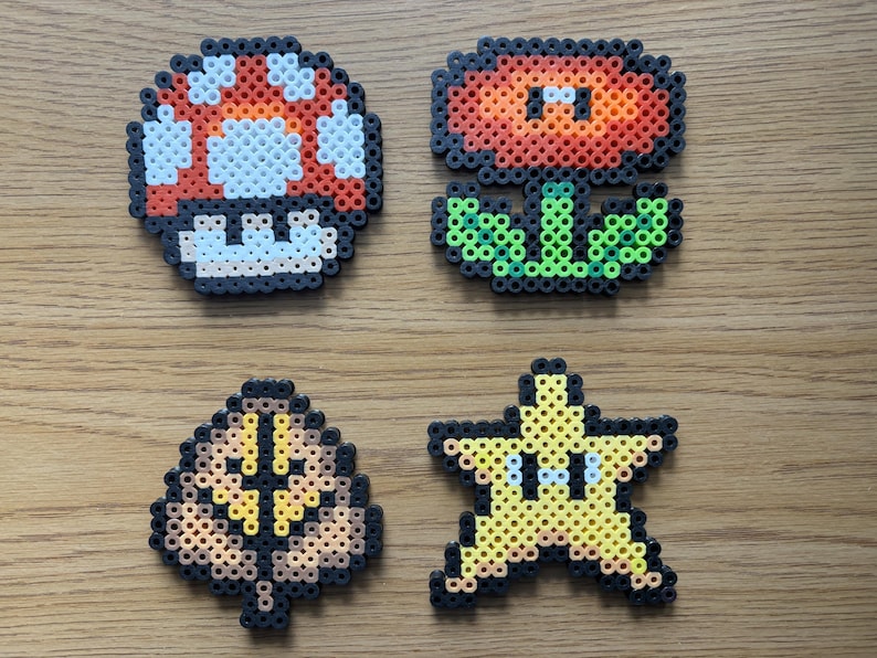 May include: Four pixel art designs from the Super Mario video game series. The designs include a red and white mushroom, a red and orange flower, a brown and yellow leaf, and a yellow star with eyes.