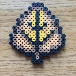 May include: A leaf-shaped bead art piece in shades of black, brown, and yellow. The design features a central yellow cross shape, surrounded by brown and black beads. The piece is made of small, fused plastic beads.