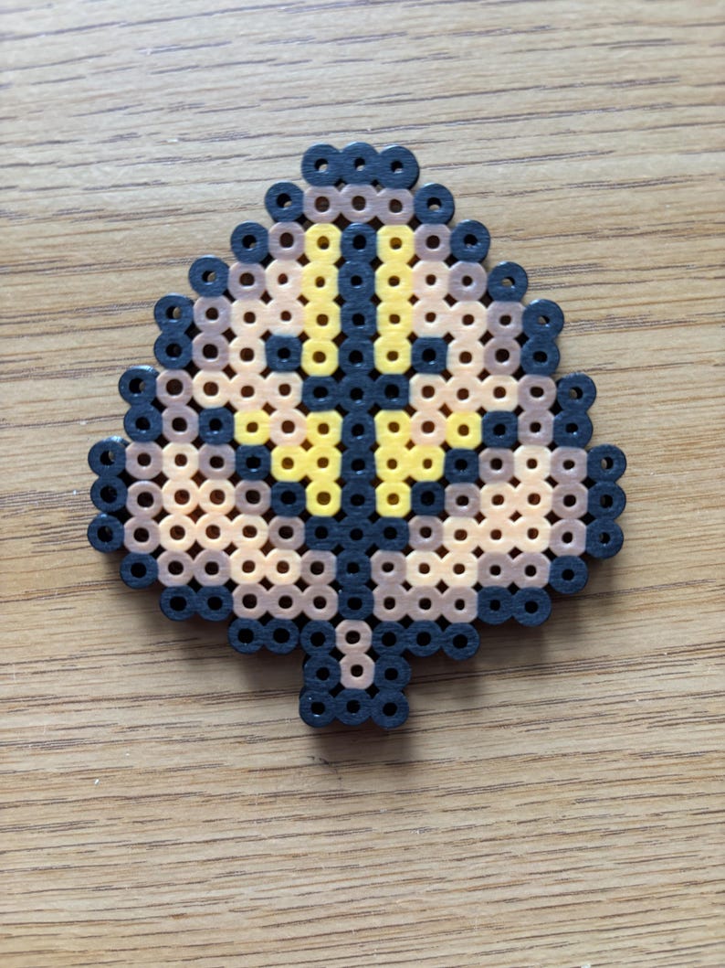 May include: A leaf-shaped bead art piece in shades of black, brown, and yellow. The design features a central yellow cross shape, surrounded by brown and black beads. The piece is made of small, fused plastic beads.