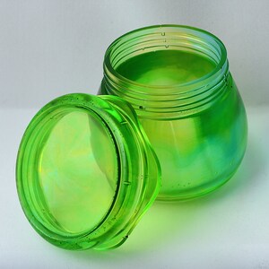 May include: A clear, iridescent green glass jar with a matching lid. The jar is round with a wide opening and a slightly tapered body. The lid is off to the side, revealing the inside of the jar. The glass has a subtle, swirling pattern.