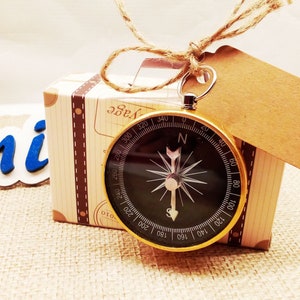 Pode incluir: Uma bússola com borda dourada e mostrador preto pende de uma pequena caixa de presente decorada como uma mala vintage. Uma etiqueta marrom em branco está anexada. A caixa é amarrada com barbante e repousa sobre uma superfície de serapilheira.