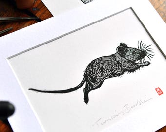 May include: Black and white linocut print of a mouse in mid-leap, set in a white mat. The artwork is signed and titled "Timian's Beanstalk". Additional linocut prints and tools are visible in the background.