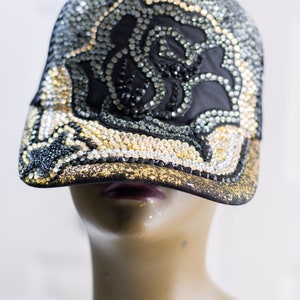 May include: A black baseball cap with a gold and silver rhinestone design featuring a rose. The cap has a curved brim and is decorated with rhinestones.