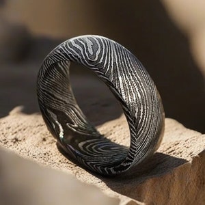 May include: A black and silver patterned ring made of metal. The ring is sitting on a light brown rock.