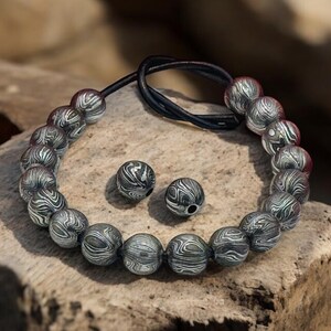May include: A beaded bracelet and two matching beads rest on a wooden surface. The bracelet features silver-colored, patterned beads strung on a black cord. The beads have a swirling design. The background is a blurred, neutral tone.