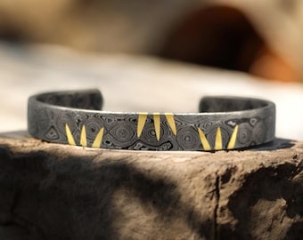 May include: A dark gray metal cuff bracelet with a patterned design and gold-colored accents. The bracelet has a series of gold-colored triangular shapes along the top. The bracelet is resting on a stone surface.
