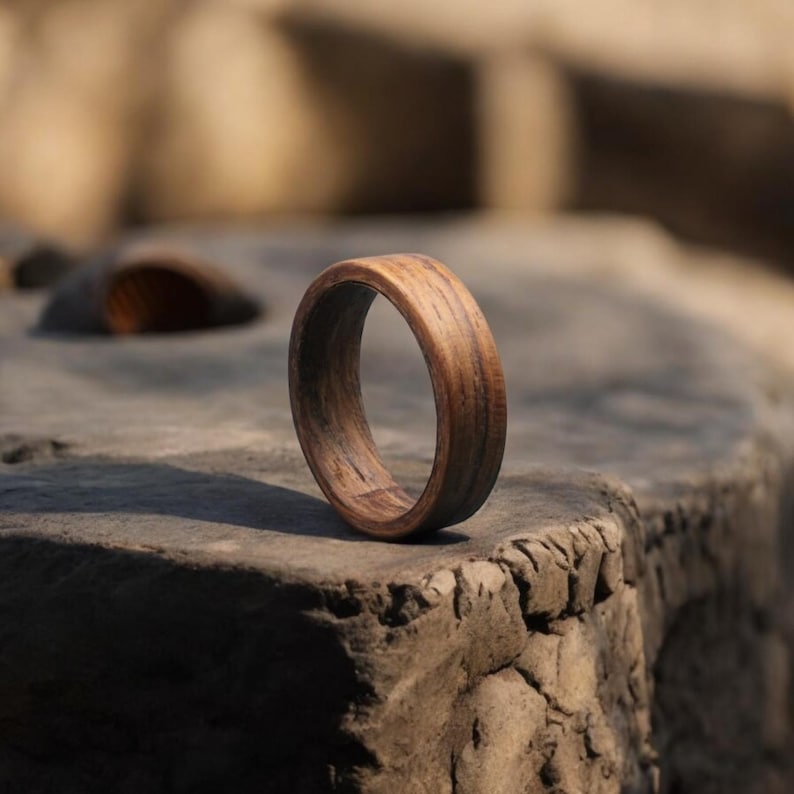 May include: A wooden ring with a natural wood grain pattern. The ring is a warm brown color and appears to be handcrafted. The ring is displayed on a textured stone surface, showcasing its organic design. This ring is a unique piece of jewelry.