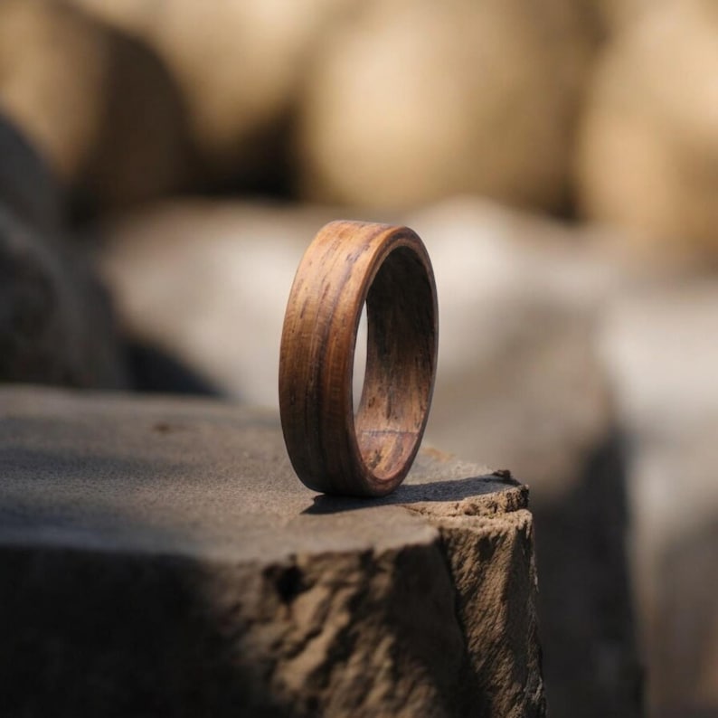 May include: A wooden ring with a natural wood grain pattern stands upright on a stone surface. The ring is a warm brown color, showcasing the wood's texture. The ring's design is simple and elegant, suitable for everyday wear.