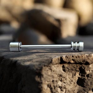 May include: A silver barbell-shaped body piercing jewelry piece. The jewelry has a cylindrical bar with a rounded end on one side and a decorative, ridged end on the other. The piece is displayed on a rough, brown surface.