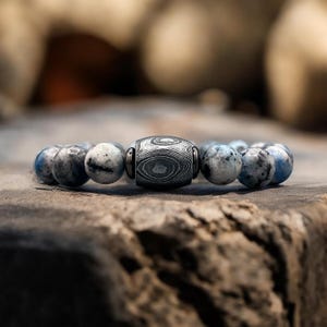 May include: A bracelet with a central patterned bead and smaller, round beads in blue, grey, and white. The central bead has a distinctive swirling pattern. The bracelet is displayed on a textured, brown surface.