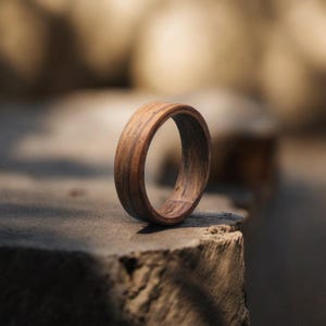 May include: A wooden ring with a natural wood grain pattern. The ring is a warm brown color and appears to be handcrafted. The ring is sitting on a stone surface, with a blurred background.