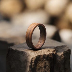 May include: A wooden ring with a natural wood grain pattern. The ring is a warm brown color and appears to be handcrafted. The ring is resting on a stone surface, showcasing its texture and design.