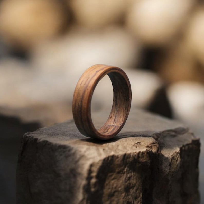 May include: A wooden ring with a natural wood grain pattern. The ring is a warm brown color and appears to be handcrafted. The ring is resting on a stone surface, showcasing its texture and design.