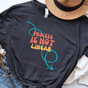 Könnte beinhalten: Ein dunkelgraues T-Shirt mit dem Text "Process is not linear" in bunten Buchstaben. Der Text ist von einer türkisfarbenen Linie umgeben, die einen Kreis mit einem Pfeil bildet.