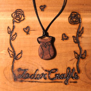 Puede incluir: Un colgante de madera con forma de búho en un collar de cordón negro. El colgante está tallado con detalles y tiene un patrón de veta de madera. El collar se muestra sobre una superficie de madera con un diseño quemado de rosas, corazones y el texto "Zodor Crafts".
