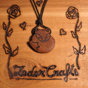 Puede incluir: Un collar con un colgante de madera en forma de zorro, con un cordón negro. El colgante está tallado con un diseño de zorro detallado y tiene un acabado de madera natural. El collar se muestra sobre una superficie de madera con un diseño de rosas y corazones quemados.