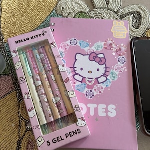 May include: A pink spiral-bound notebook featuring a Hello Kitty graphic surrounded by a heart of colorful gems and the text NOTES, paired with a box of five Hello Kitty gel pens in pastel shades, each decorated with small icons of bows and bags.