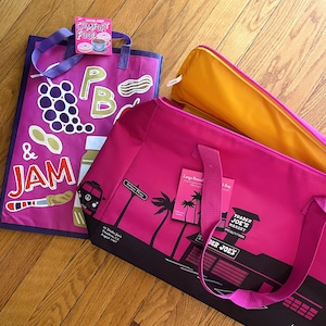 May include: Three reusable shopping bags on a wooden floor. One is magenta with a grape and jam design, another is pink with a palm tree graphic, and the third is yellow. A small box with the words "COMFORT FOOD" is on top of the magenta bag.