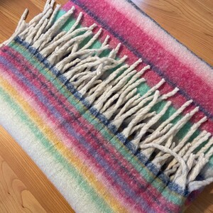 Op de afbeelding: Een veelkleurige deken met een gestreept patroon in roze, blauw, groen en geel. De deken heeft een franje van witte kwasten langs &eacute;&eacute;n rand. De deken is opgevouwen en ziet er zacht en gezellig uit.