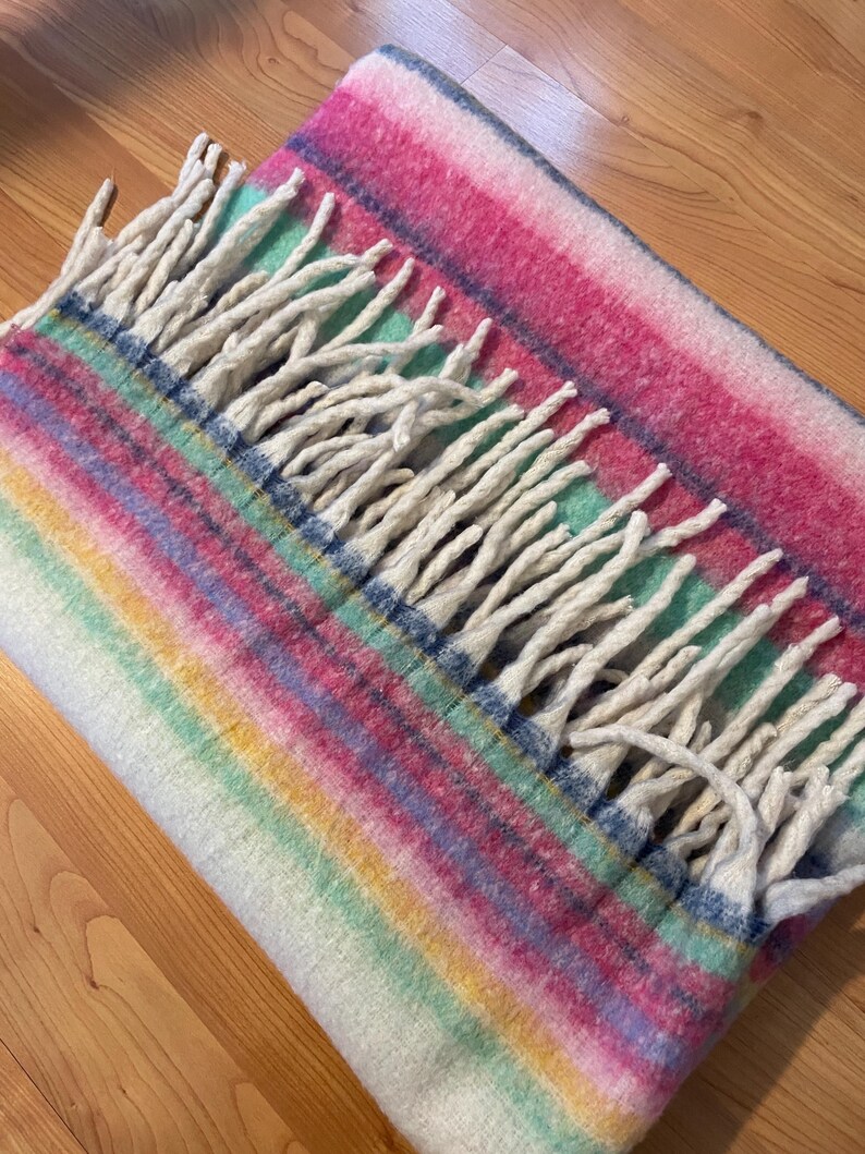 Op de afbeelding: Een veelkleurige deken met een gestreept patroon in roze, blauw, groen en geel. De deken heeft een franje van witte kwasten langs &eacute;&eacute;n rand. De deken is opgevouwen en ziet er zacht en gezellig uit.