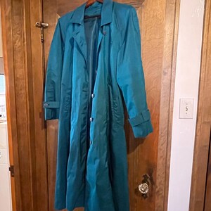 May include: A teal-colored trench coat hangs on a wooden door. The coat has a classic design with a collar, buttons, and long sleeves. The coat is made of a smooth, possibly water-resistant material. The coat is a long length, reaching near the ankles.