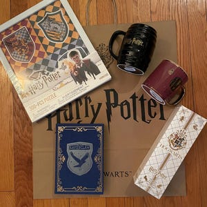 May include: A collection of Harry Potter merchandise including a puzzle, mugs, a book, and a box. The puzzle features the Hogwarts crests and Harry Potter. The mugs are black and burgundy. The book is blue with a Ravenclaw crest.