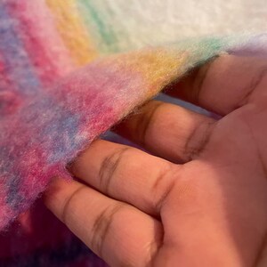 Op de afbeelding: Close-up van een zachte, veelkleurige stof met een regenboogverloop. De kleuren zijn roze, blauw, geel en groen. De stof lijkt pluizig en getextureerd, met een witte achtergrond.