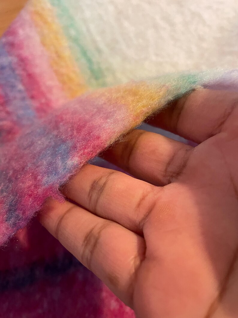Op de afbeelding: Close-up van een zachte, veelkleurige stof met een regenboogverloop. De kleuren zijn roze, blauw, geel en groen. De stof lijkt pluizig en getextureerd, met een witte achtergrond.