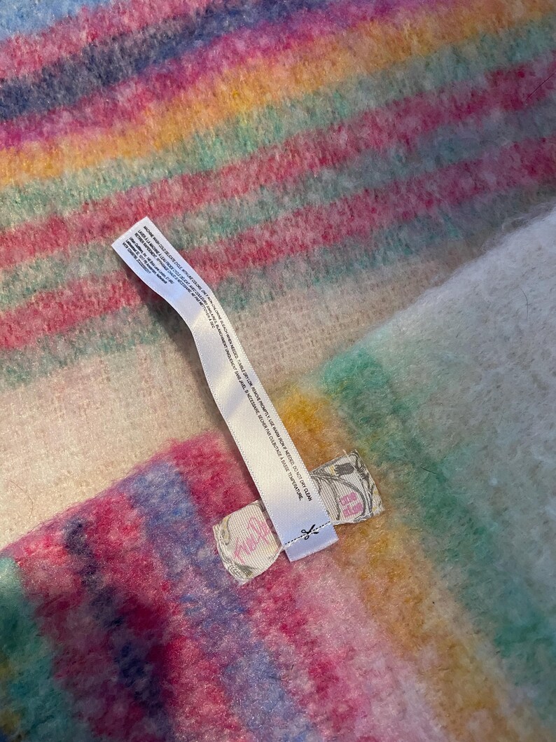 Op de afbeelding: Een veelkleurig gestreept textiel in roze, blauw, geel en groen. Een wit label met tekst is aan het textiel bevestigd. Het label heeft een kleine illustratie en het woord "Free People".