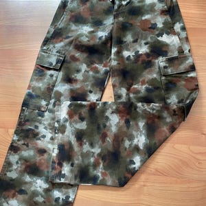 May include: Camouflage cargo pants with a brown, green, and white color scheme. The pants have a relaxed fit and two large cargo pockets on the sides.