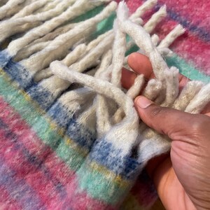 Op de afbeelding: Close-up van een kleurrijke geweven deken met een roze, blauw, groen en geel geruit patroon. De deken heeft lange, cr&egrave;mekleurige franjes. De textuur van de deken is zacht en uitnodigend.