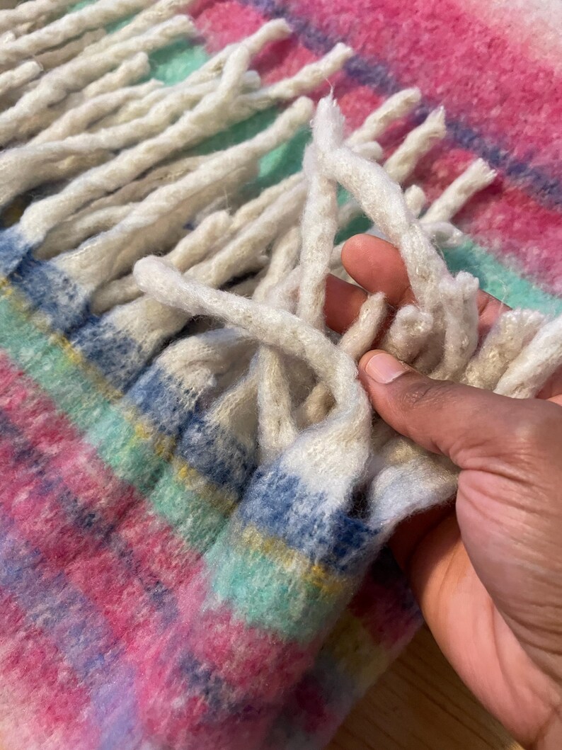 Op de afbeelding: Close-up van een kleurrijke geweven deken met een roze, blauw, groen en geel geruit patroon. De deken heeft lange, cr&egrave;mekleurige franjes. De textuur van de deken is zacht en uitnodigend.