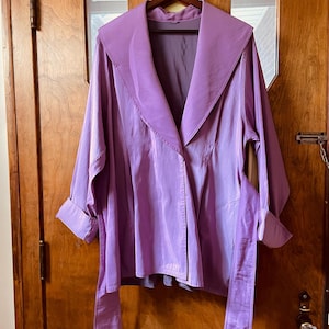 May include: A lilac-colored jacket with a wide collar and long sleeves hangs on a wooden hanger. The jacket features a tie belt and is made of a shiny, silky material. The cuffs are rolled up, and the jacket is displayed against a wooden door.