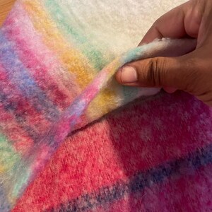 Op de afbeelding: Een close-up van een zachte, kleurrijke deken met een regenboogverloop van roze, blauwe, gele en groene strepen. De deken heeft een zachte textuur en een witte achtergrond.