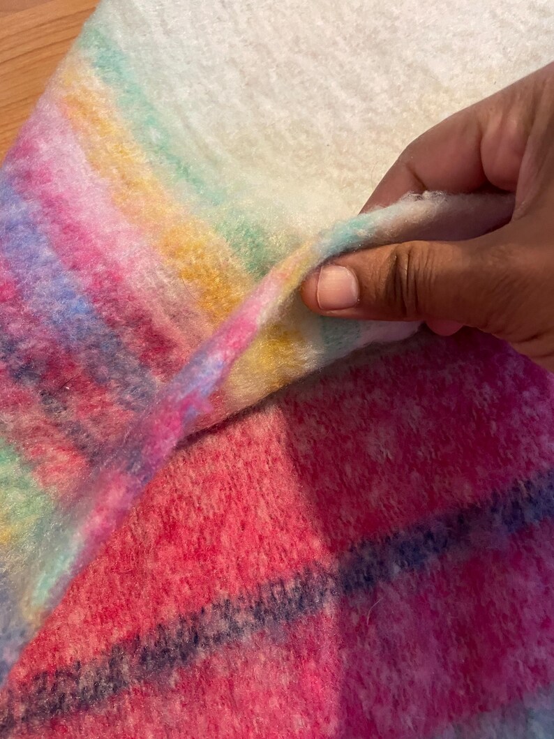 Op de afbeelding: Een close-up van een zachte, kleurrijke deken met een regenboogverloop van roze, blauwe, gele en groene strepen. De deken heeft een zachte textuur en een witte achtergrond.