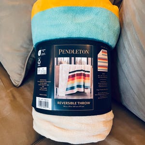 May include: A rolled-up reversible throw blanket. The top is yellow, turquoise, and white. The label reads "PENDLETON REVERSIBLE THROW" and lists dimensions as 50 in. x 70 in. (127 cm x 177 cm). The blanket is on a brown surface.