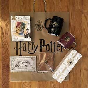 May include: A collection of Harry Potter themed merchandise on a brown paper shopping bag. Items include a puzzle, two mugs, a journal, a wand box, and a ticket. The bag and items feature the Harry Potter logo and Hogwarts crest.