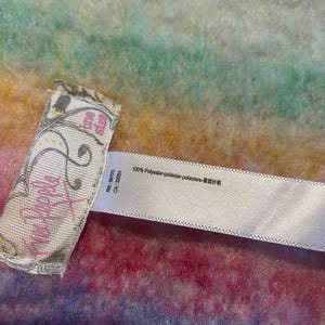 Op de afbeelding: Close-up van een label van Free People kleding. Het label is wit met roze tekst en decoratieve krullen. De achtergrond is een kleurrijke, getextureerde stof met tinten roze, blauw, groen en geel. Het label bevat ook de tekst "100% Polyester".