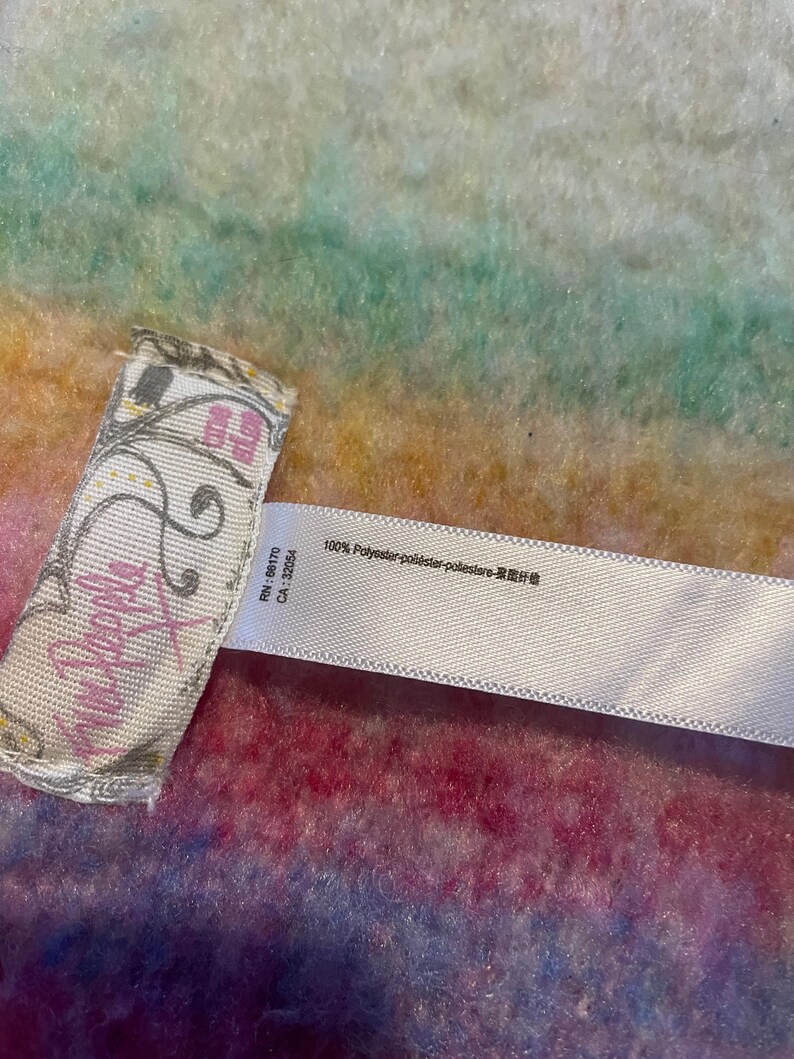 Op de afbeelding: Close-up van een label van Free People kleding. Het label is wit met roze tekst en decoratieve krullen. De achtergrond is een kleurrijke, getextureerde stof met tinten roze, blauw, groen en geel. Het label bevat ook de tekst "100% Polyester".