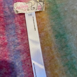 Op de afbeelding: Een kleurrijk, getextureerd textiel met een label met de tekst "Free People" en "one size". De stof heeft een mix van roze, blauwe, gele en groene tinten. Een wit label met wasvoorschriften is ook zichtbaar.