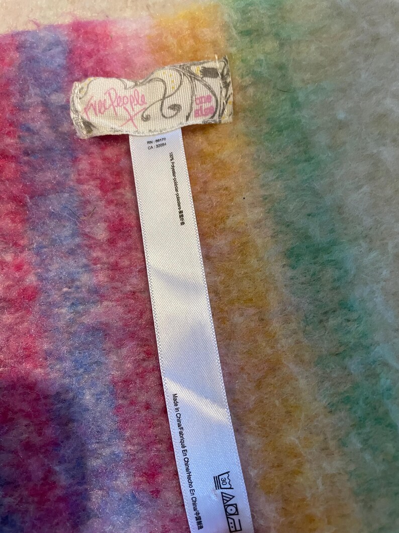 Op de afbeelding: Een kleurrijk, getextureerd textiel met een label met de tekst "Free People" en "one size". De stof heeft een mix van roze, blauwe, gele en groene tinten. Een wit label met wasvoorschriften is ook zichtbaar.