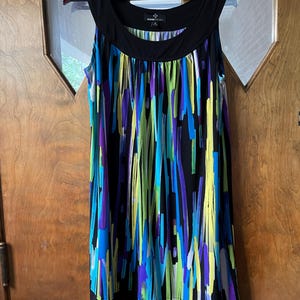 May include: Sleeveless dress with a black neckline and hem. The dress features a vibrant pattern of vertical stripes in blue, purple, and green against a black background. The dress is hanging on a wooden hanger.