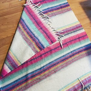 Op de afbeelding: Een kleurrijke gestreepte deken met roze, blauwe, groene en gele banden op een witte achtergrond. De deken heeft een gefranjerde rand. Perfect om een vleugje kleur aan een kamer toe te voegen.