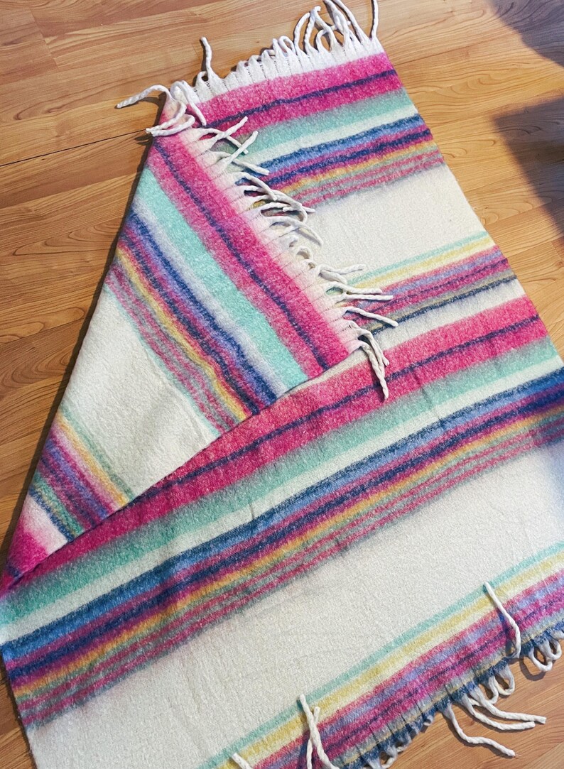 Op de afbeelding: Een kleurrijke gestreepte deken met roze, blauwe, groene en gele banden op een witte achtergrond. De deken heeft een gefranjerde rand. Perfect om een vleugje kleur aan een kamer toe te voegen.