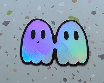 May include: A holographic sticker featuring two cartoon ghosts. The ghosts are side-by-side, with a black outline. One ghost has a purple hue, and the other has a green and blue hue. Each ghost has two black oval eyes.