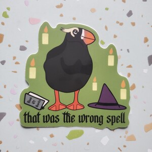 May include: A green sticker with a black and white illustration of a puffin wearing a witch's hat. The puffin is standing next to a book that says "Book of Spells" and a few candles. The text "that was the wrong spell" is written below the puffin.