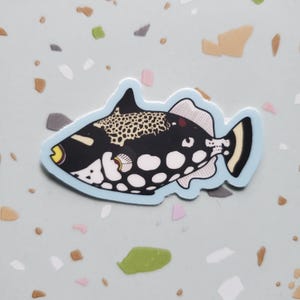May include: A sticker of a black and white triggerfish with a yellow mouth and a blue outline. The fish has a spotted pattern on its back.