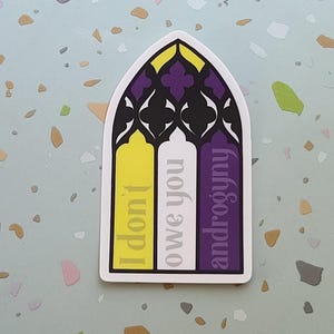 May include: A sticker in the shape of a stained-glass window with the words "I don't owe you androgyny." The sticker features yellow, white, and purple vertical stripes, with black and purple decorative elements at the top.