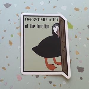 May include: A sticker featuring a cartoon puffin peeking out from behind a door. The puffin is black with a white face and orange feet. The text "OVERSTIMULATED at the function" is above the bird.