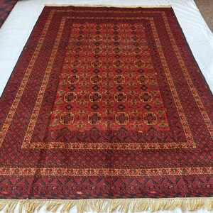 May include: A rectangular area rug featuring a deep red field with repeating dark geometric motifs and octagonal patterns. The rug is framed by multiple intricate borders in shades of black, red, and gold, finished with light-colored fringe.